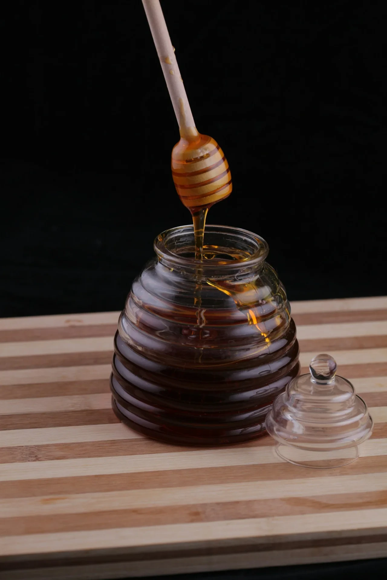 Golden honey dripping slowly from a wooden spoon over a glass jar, warm amber tones, natural light from window